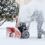 man snowblowing a sidewalk with a tree behind him and a yellow building