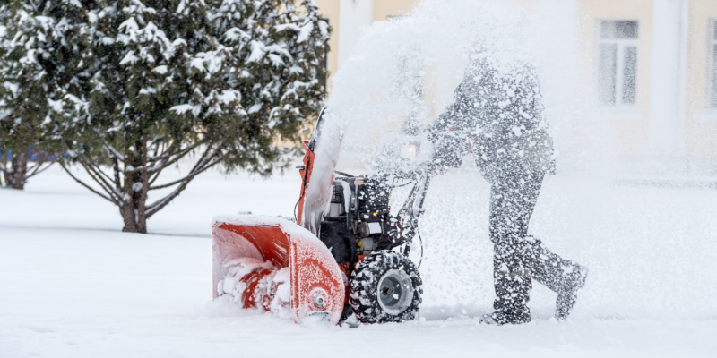 man snowblowing a sidewalk with a tree behind him and a yellow building