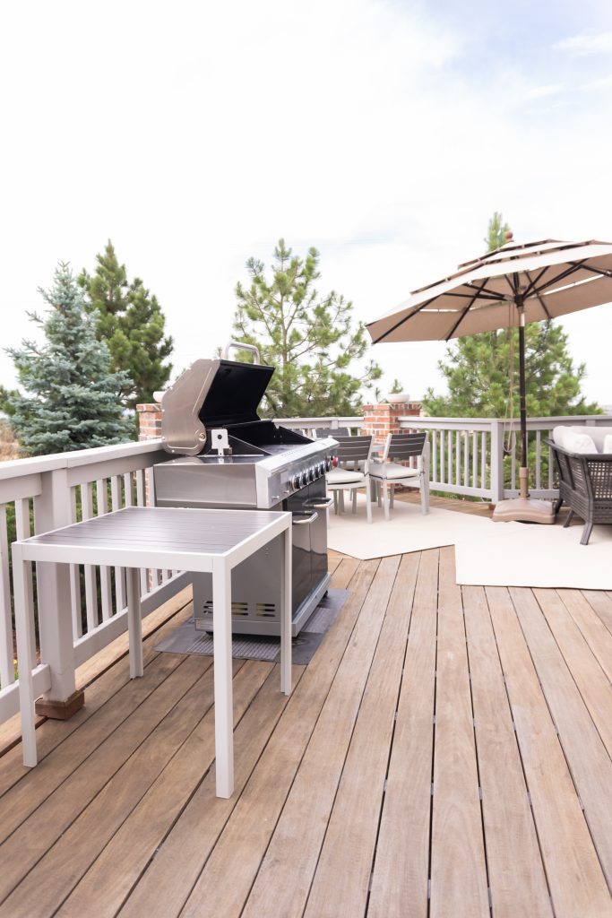 A silver-colored barbecue grill sits on a wooden deck. There is a small white table next to the grill.
