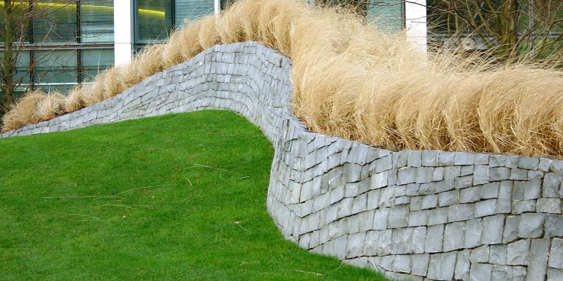 Lush green grass meets a stacked stone wall, topped with tan ornamental grass, acting as erosion control in Roanoke.