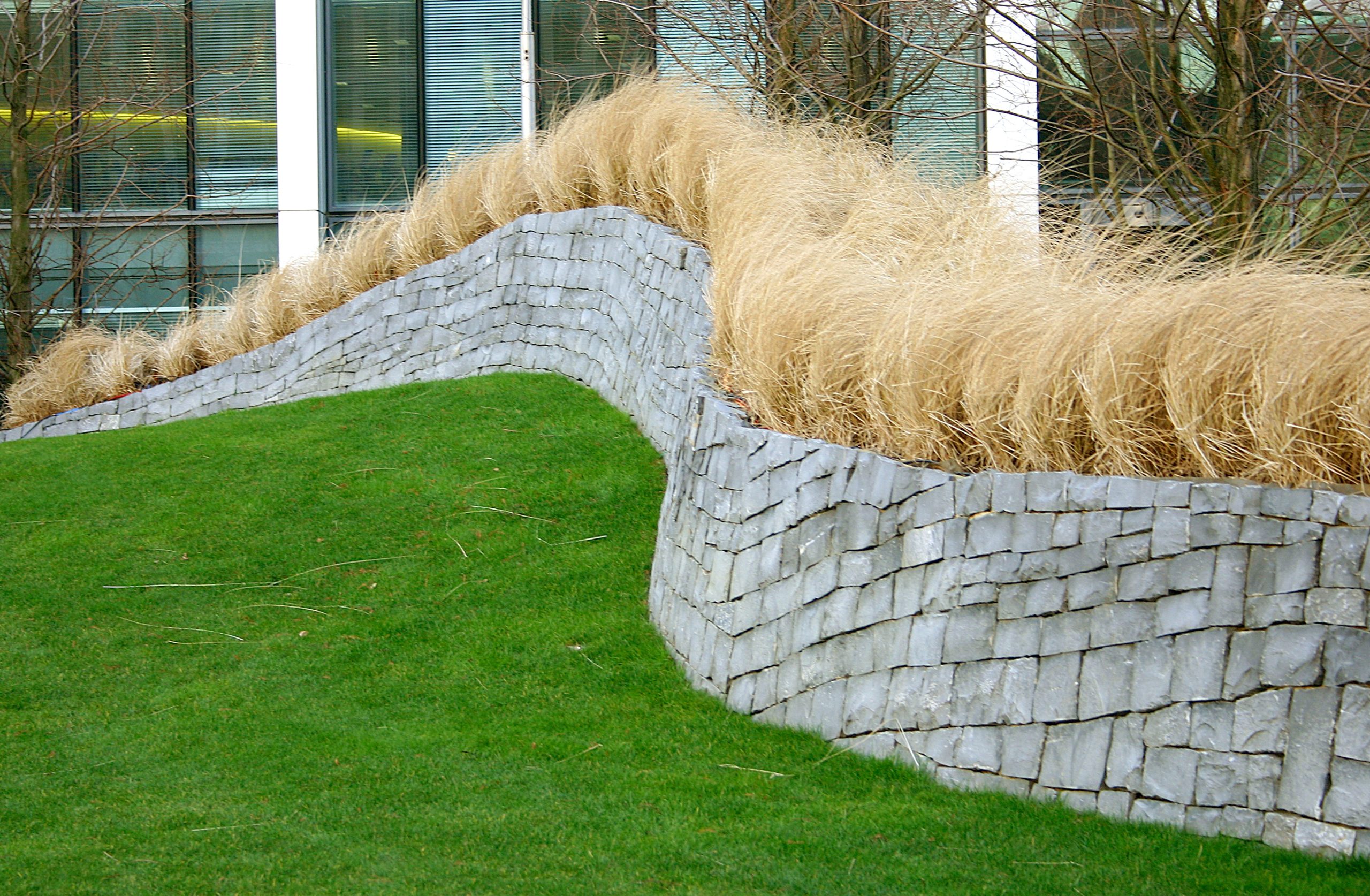 Lush green grass meets a stacked stone wall, topped with tan ornamental grass, acting as erosion control in Roanoke.